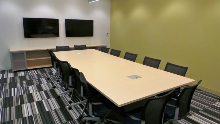 A long conference room table surrounded by chairs. There are 2 screens on the back wall of the room
