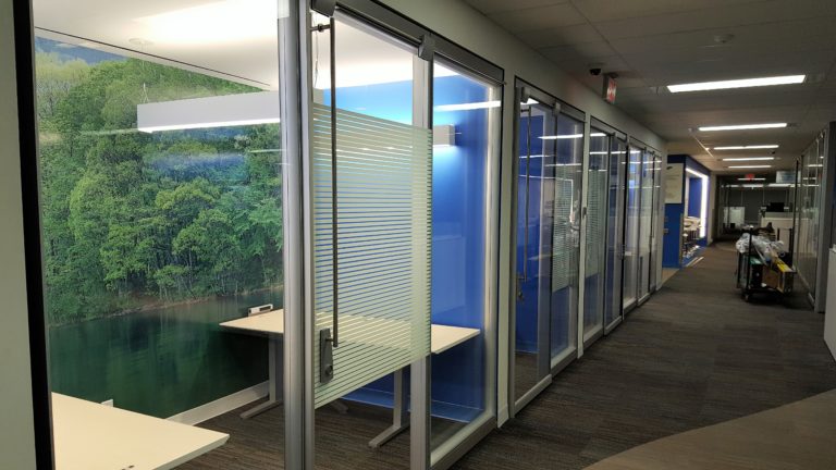 A hallway with glass walls looking into offices with blue and green walls.