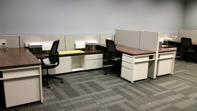 A shared cubicle with two black desk chairs, one wooden topped desk, and one table between the two desks.